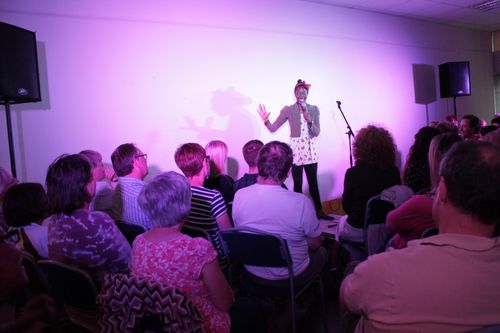 Performer speaking to an audience of people, with a Microphone in her hand. The lighting on the stage is purple, and two speakers stand at either side of the stage.